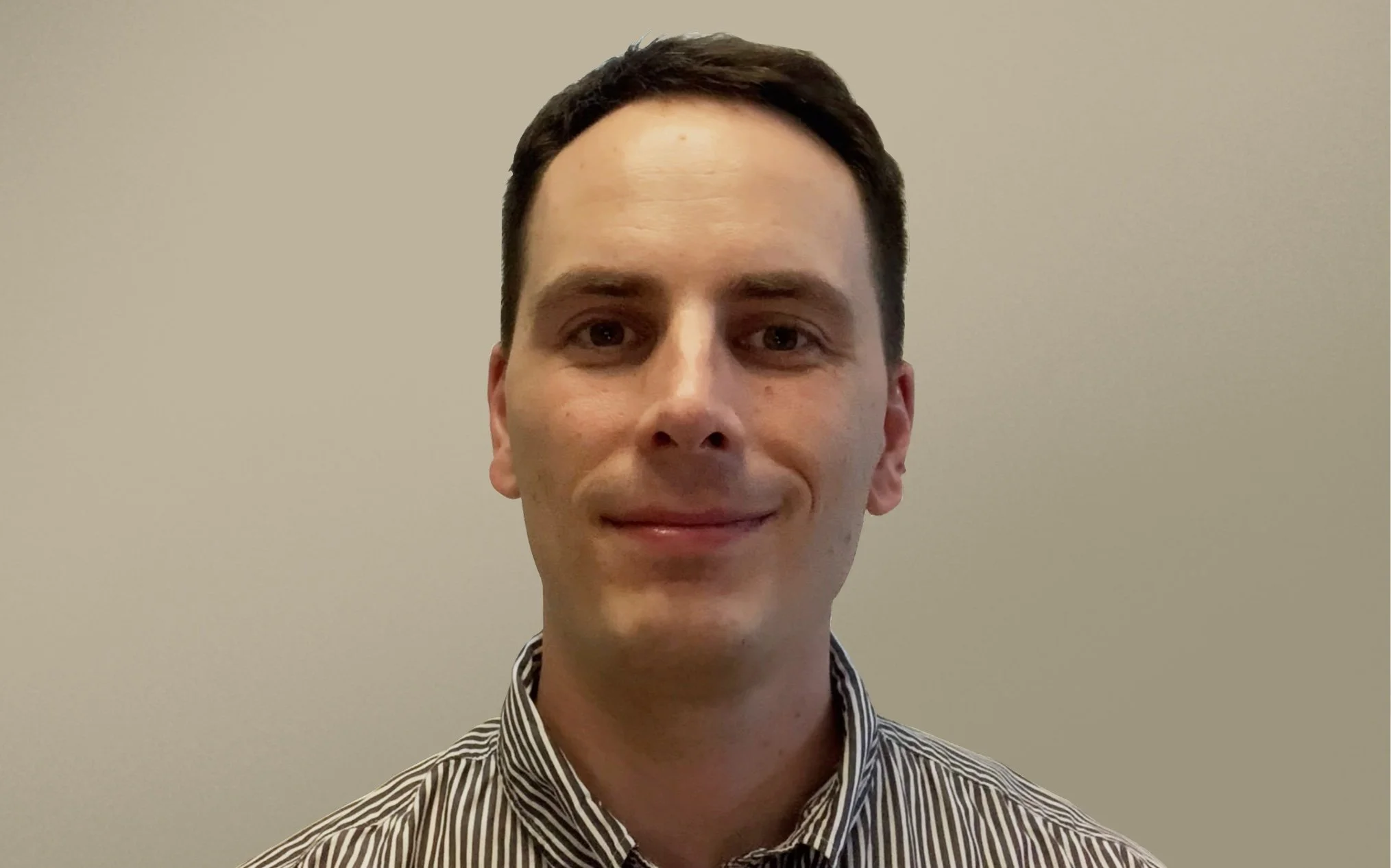 A young man with short dark hair, brown eyes, and a light complexion, smiling slightly, wearing a black and white striped shirt, against a plain beige background.