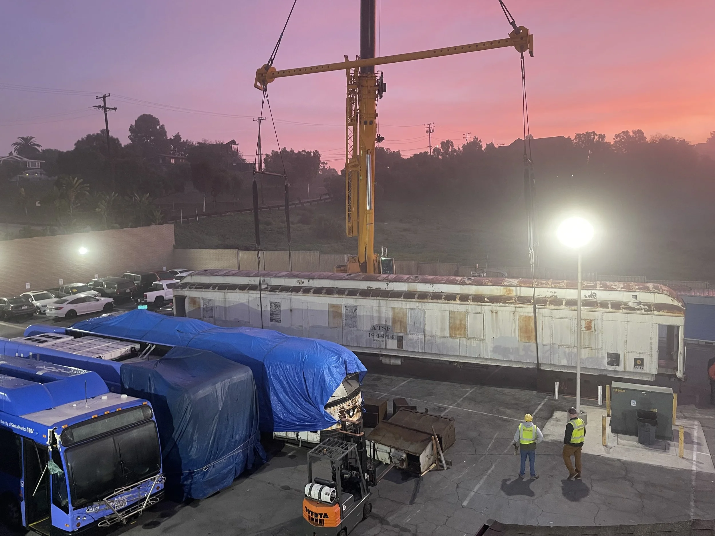 A parking lot with vehicles covered by blue tarps, a large yellow construction crane, and two workers in safety vests and hard hats, with overcast sky and power lines in the background.