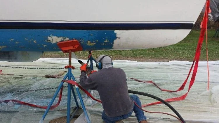 A person working on the underside of a boat, which is propped up on boat stands, with tools and equipment around.