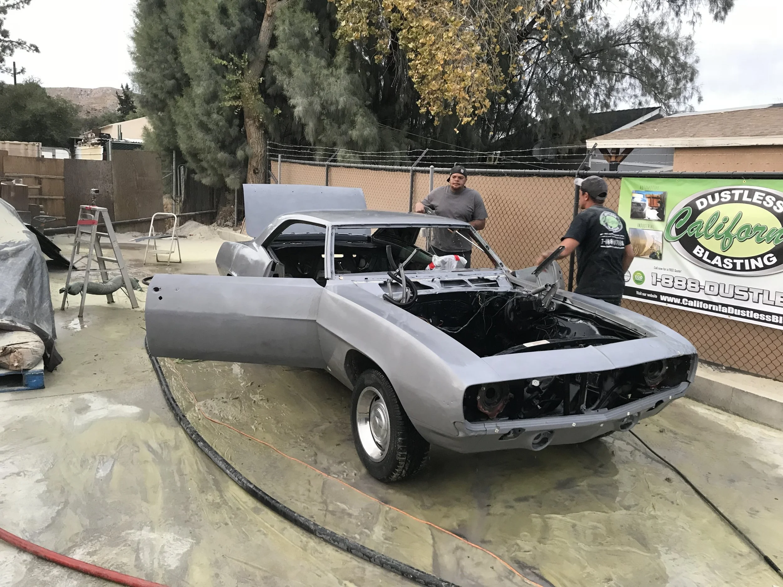 A partially assembled classic car inside a garage or workshop, with two men working on it. The car is metallic gray, and its hood is open, exposing the engine compartment. Surrounding the car are tools, equipment, a green banner for California Dustless Blasting, and some trees visible through a fence in the background.