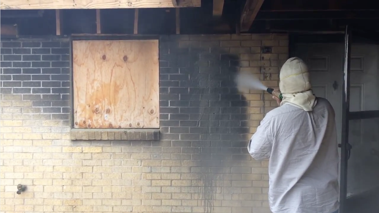 Person in protective clothing using a power washer to clean a brick wall, with a boarded-up window in background.