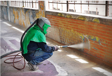 Worker wearing a helmet and green jacket pressure washing a brick wall with a hose along a sidewalk.