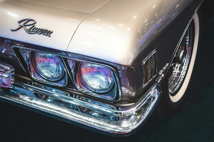 Close-up of the front left side of a vintage Oldsmobile with a chrome bumper and dual headlights, showing part of the silver body and whitewall tire.