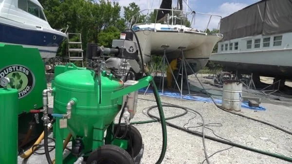 A green pressure washer cleaning boat hulls at a boatyard, with other boats and equipment in the background.