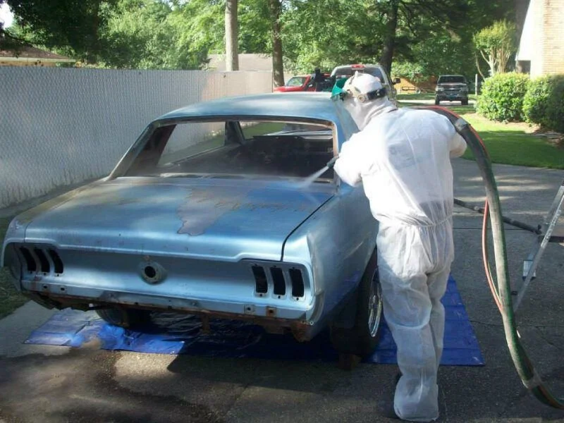 Person in protective suit spray painting a vintage car outdoors on a driveway.