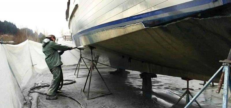 A person in green work clothes using a tool to clean or repair the hull of a boat supported by scaffolding in a boatyard.