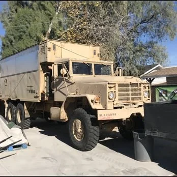 A beige military-style truck with a covered cargo area parked on a driveway, with trees and a house in the background.