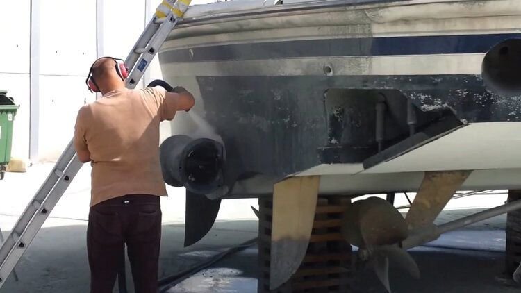 A person working on a boat, wearing headphones, using a tool near the hull, with a ladder positioned against the boat.