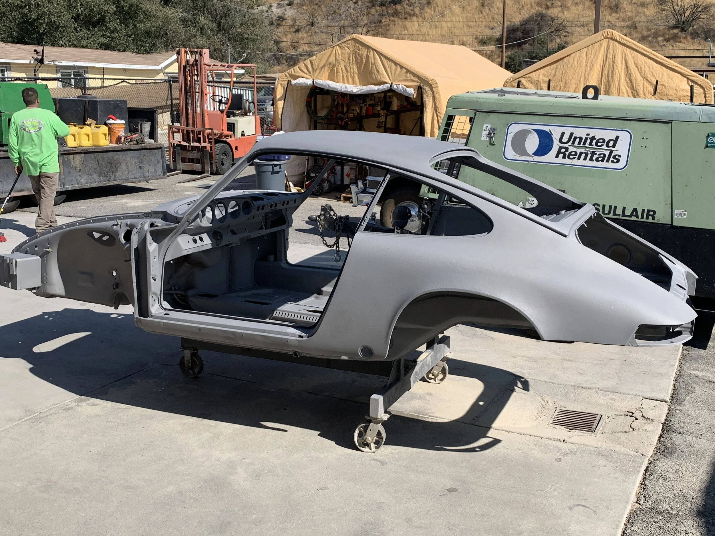 Scene of a vehicle repair or restoration shop with a partially assembled sports car, tool tents, a forklift, and various equipment and supplies.