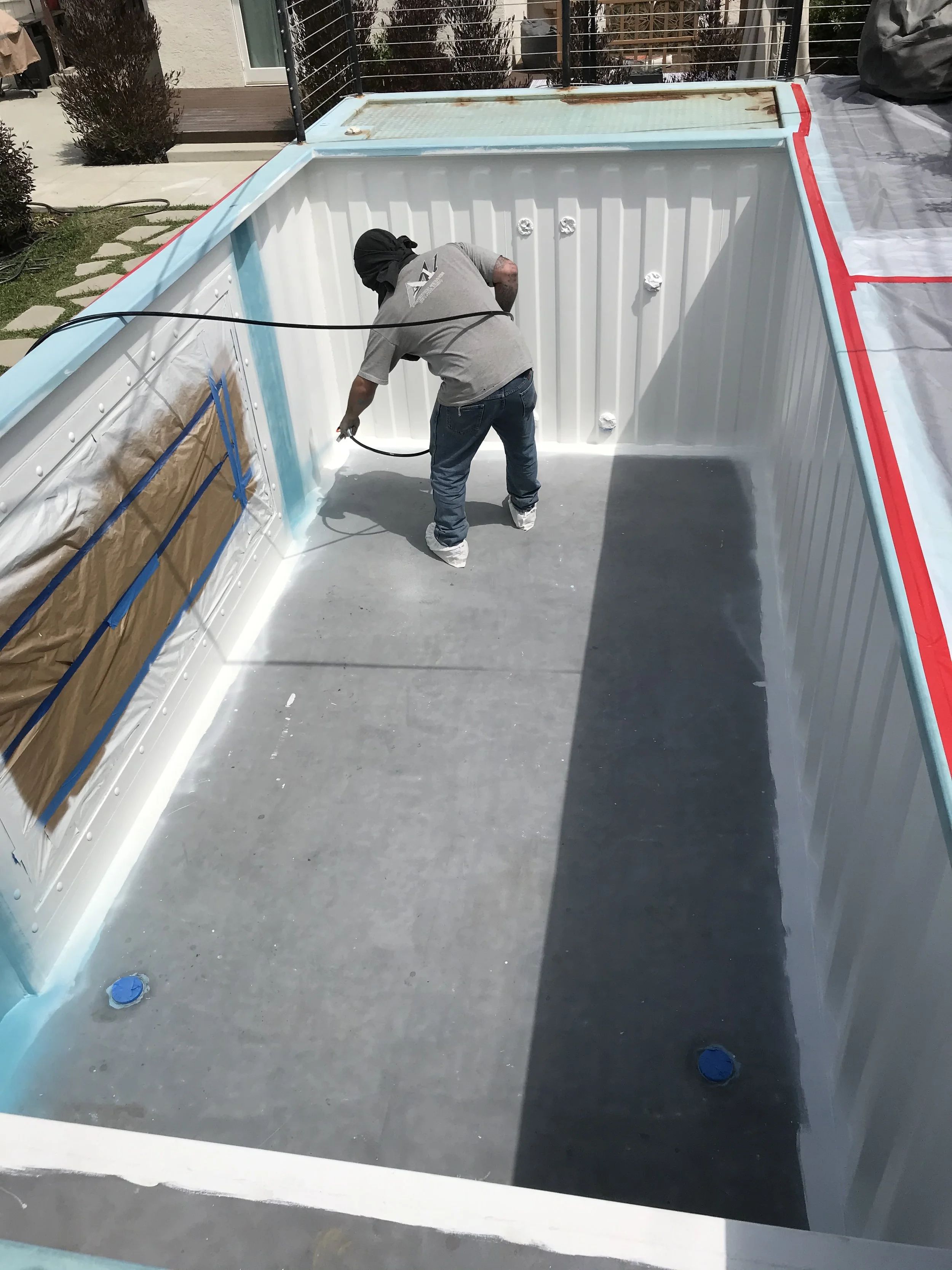 A person wearing a gray shirt, jeans, and a black head covering inside an empty swimming pool, painting the interior with a spray gun. The pool is set in a backyard with a patio, door, and outdoor plants.