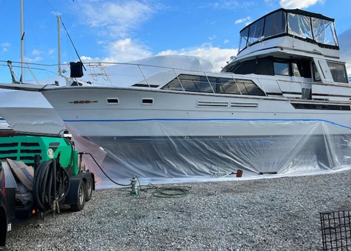 Large white motorboat on a gravel surface, covered with a clear plastic tarp, with a green and black trailer and equipment nearby, under a partly cloudy sky.