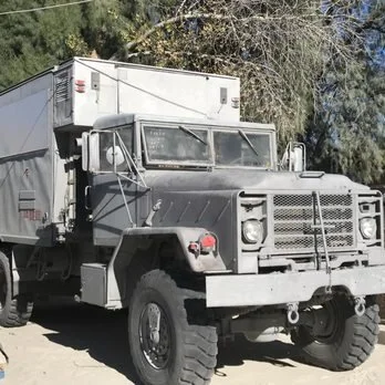 A large, gray military-style truck with a cab-over-engine design, parked outdoors near trees.