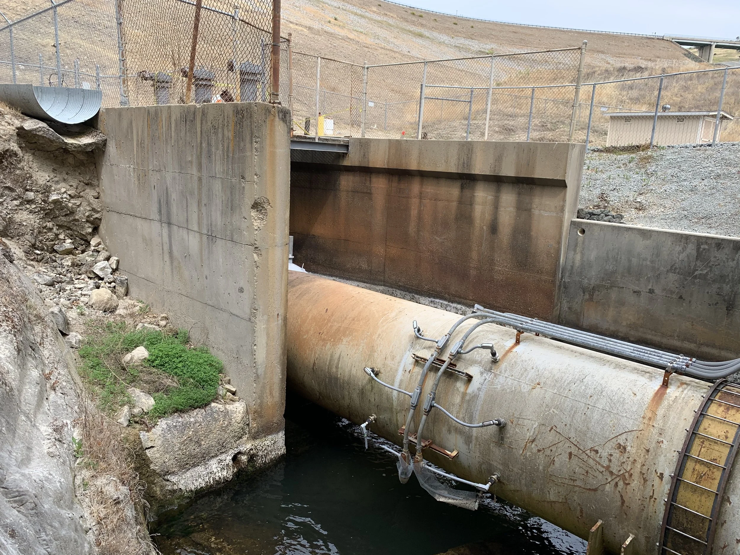 A concrete dam with water flowing underneath, surrounded by rocky terrain with some green vegetation, and chained fencing on the top of the dam, with hills in the background.
