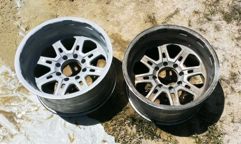Two car rims placed outdoors on the ground, one with a white finish and the other with a silver finish.