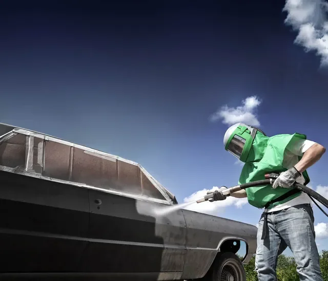 Person in a green protective suit and helmet power washing a boat outdoors under a blue sky with clouds.