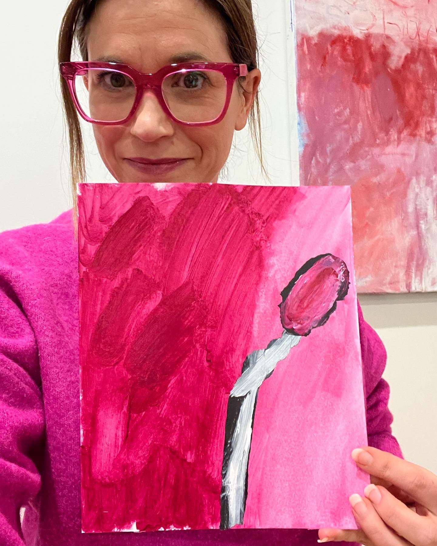 A woman with red hair, pink glasses, and a pink jacket holding a pink abstract painting of a flower with a white stem and a single pink blossom.