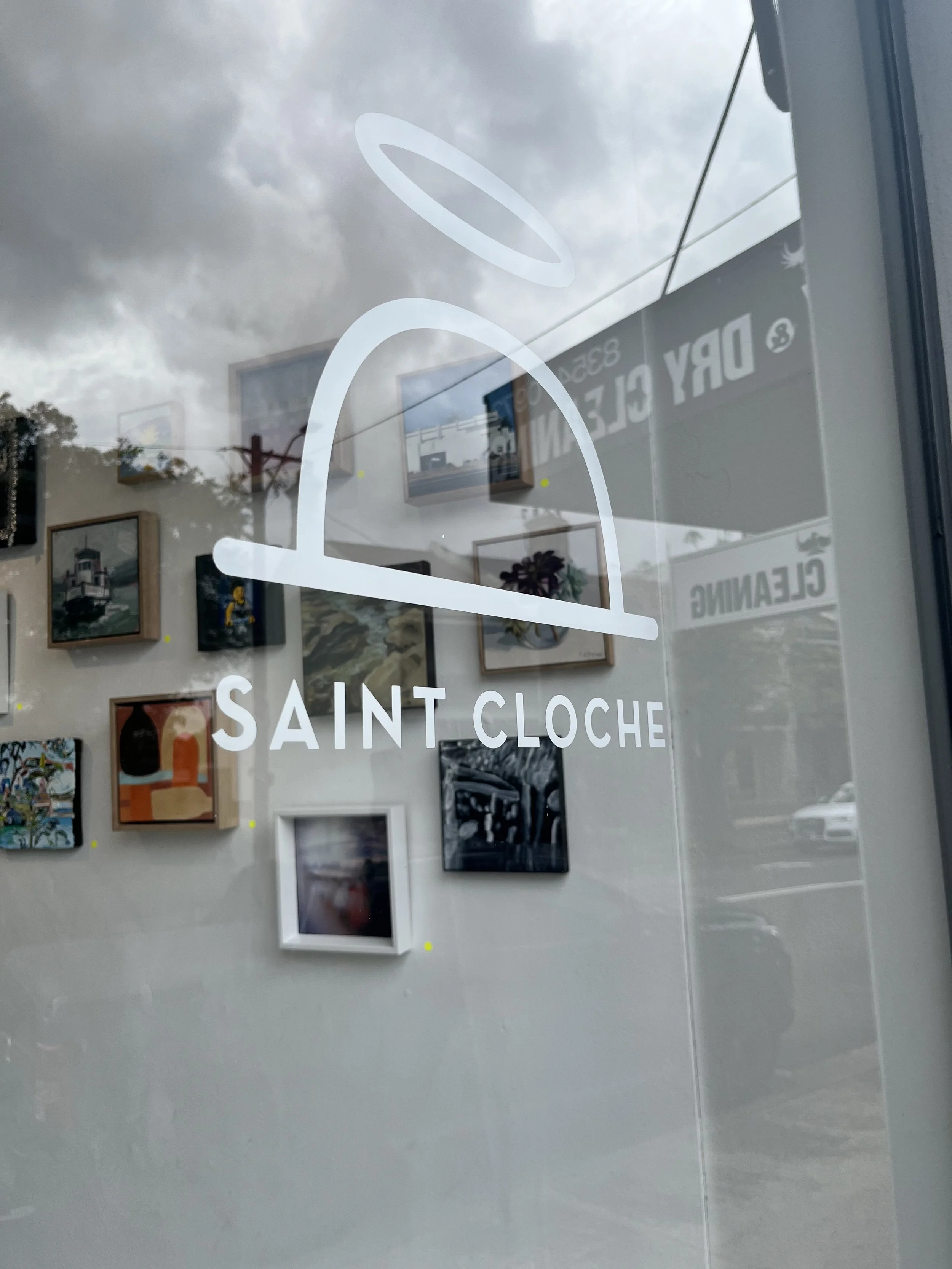 Reflection of an art gallery interior with paintings, framed art pieces on the wall, and signs for Saint Cloche and Dry Clean, seen through a glass window with a white logo