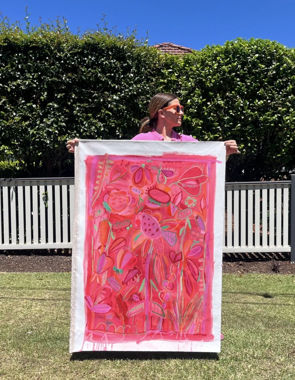 A woman in pink clothing and orange sunglasses holding a colorful pink abstract floral painting outdoors against a green hedge and a white fence, under a clear blue sky.