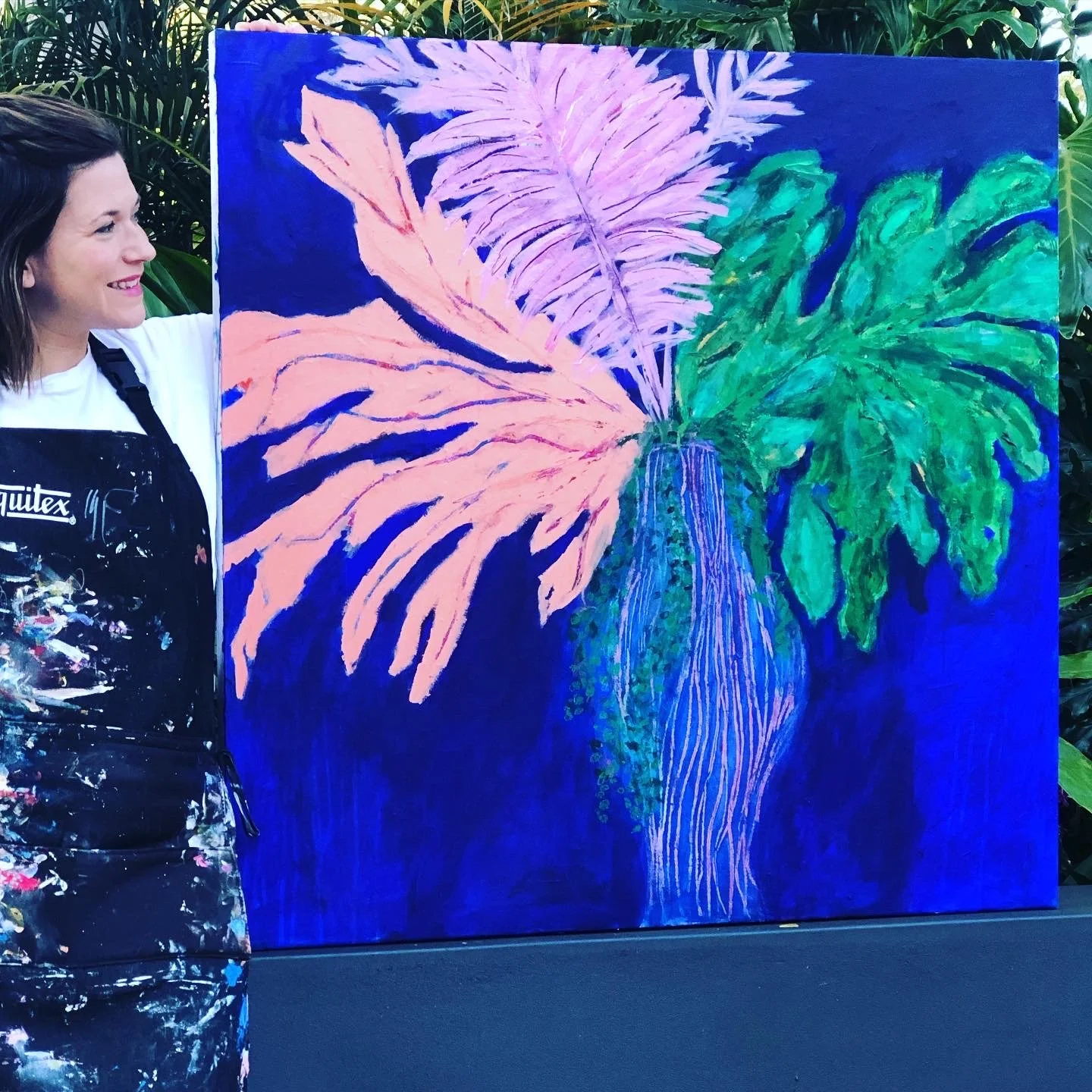 A woman standing beside a vibrant painting of large tropical leaves in pink, purple, green, and blue hues, set against a dark background.