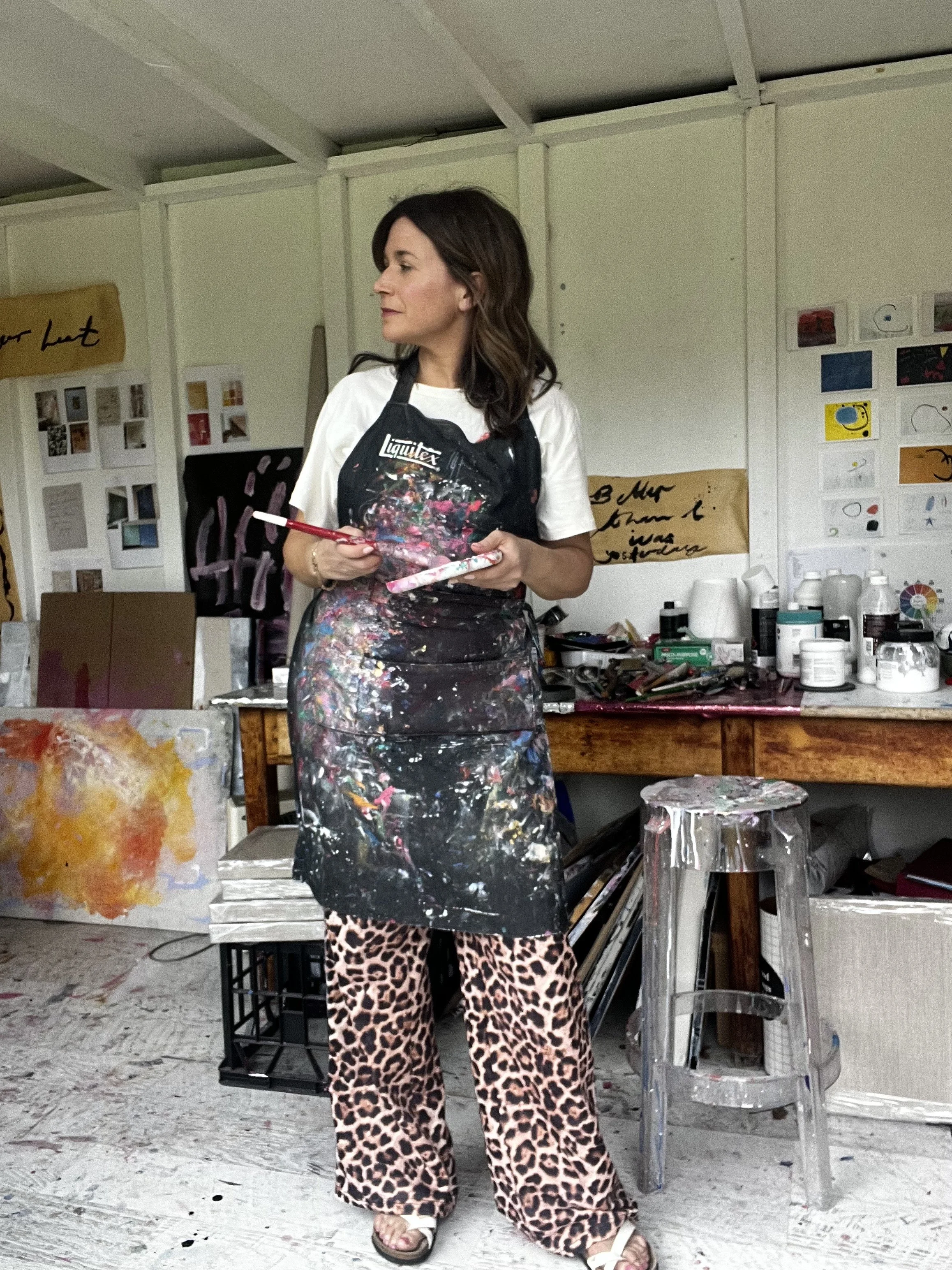 A woman stands in an art studio holding a paintbrush and palette. She is wearing a black paint-splattered apron, leopard print pants, and sandals. The background shows paintings, art supplies, and a worktable.