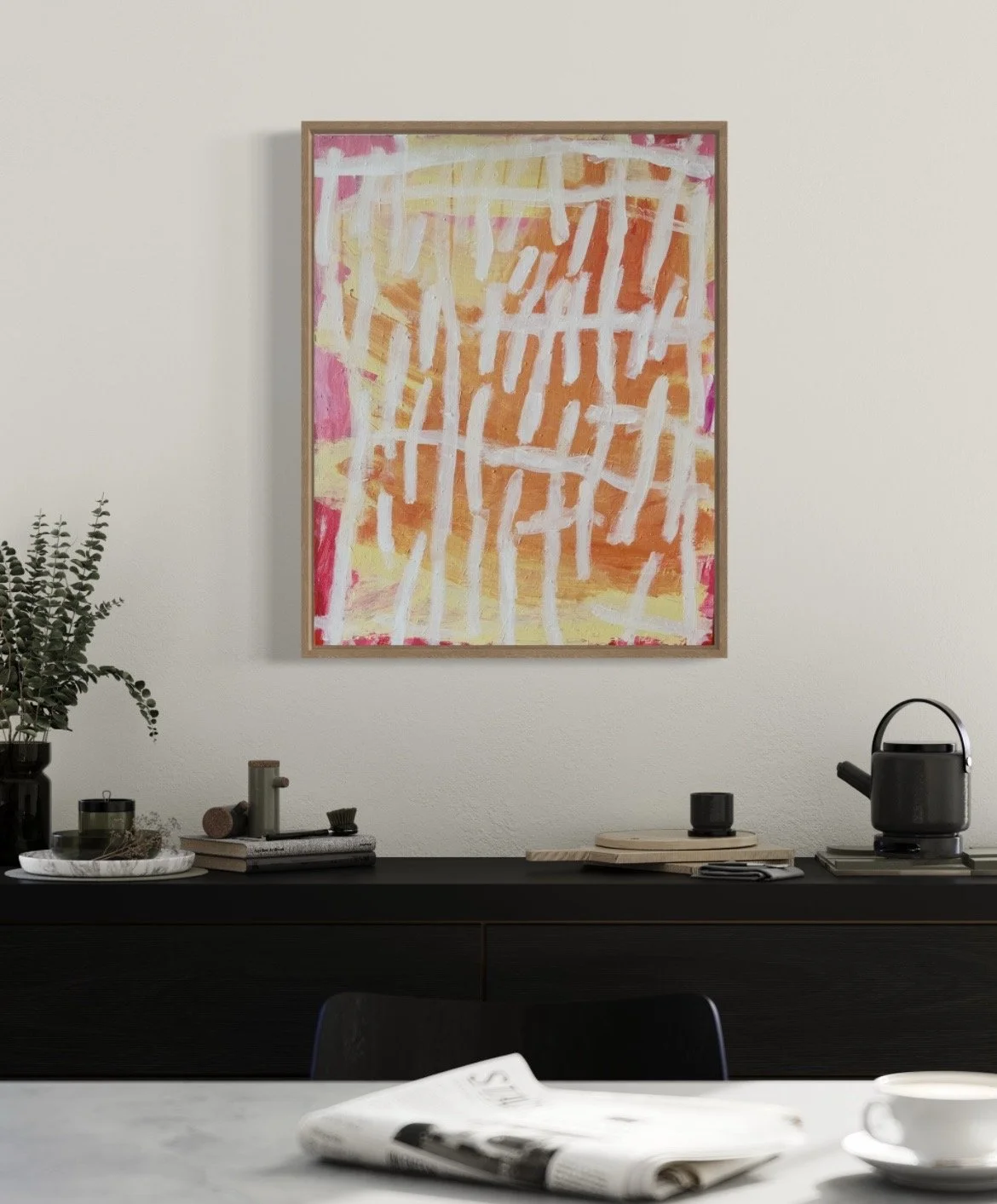 An abstract painting with pink, orange, and yellow hues hanging on a white wall above a black sideboard. The sideboard has a potted plant, a marble tray with small items, a few books, a black cup, a black teapot, and a black kettle. A magazine is on 