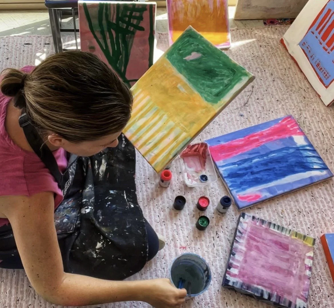 A woman painting colorful abstract art on canvas with various paint bottles and brushes on a table covered with a cloth.
