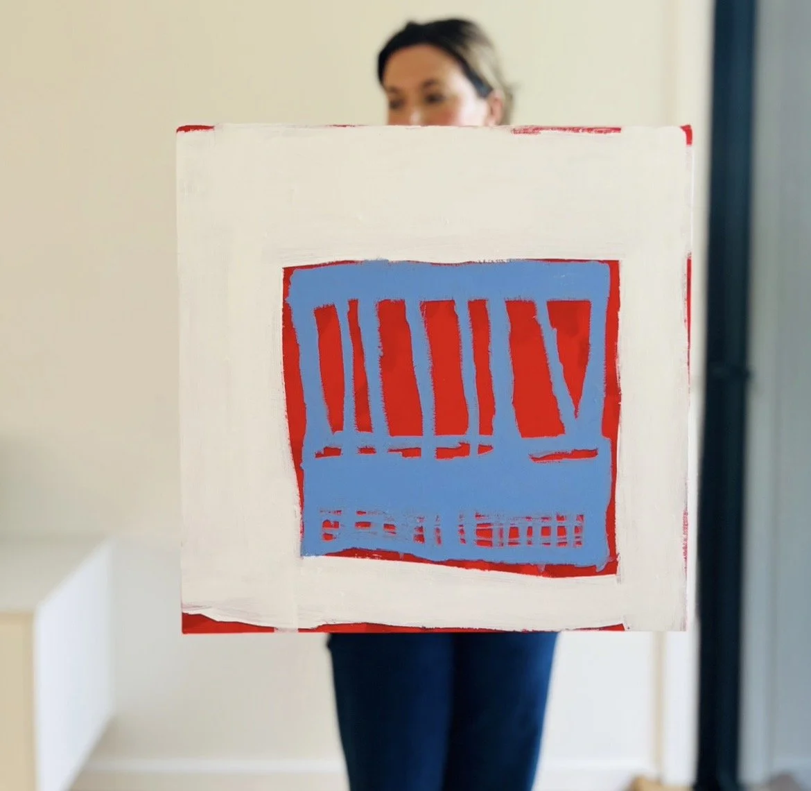 Person holding a cardboard box with a red and blue barcode design painted on it, partially obscuring their face, in a room with light-colored walls.