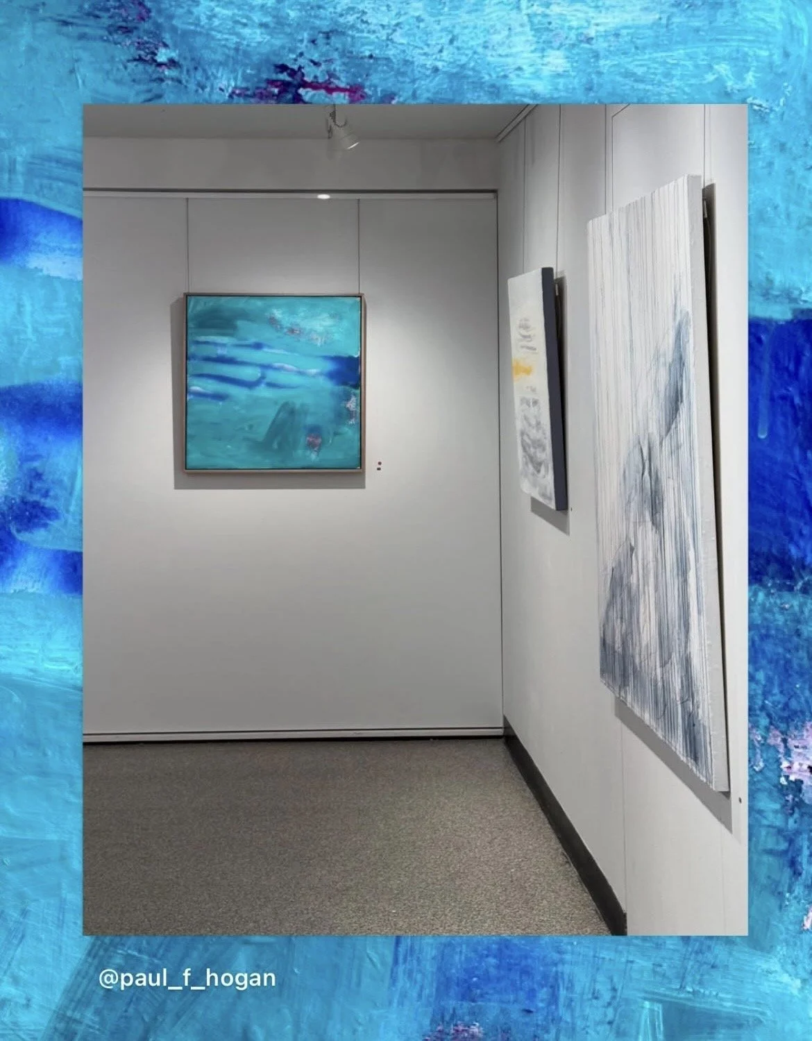 View of an art gallery with two paintings on white walls and a blue abstract painting framed on the back wall. The gallery has gray carpeted flooring and a ceiling with track lighting.