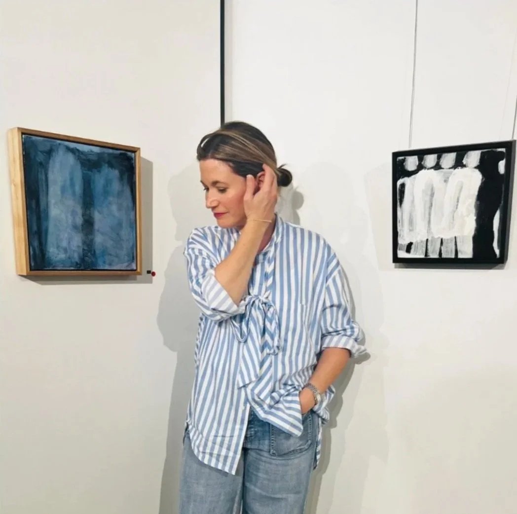 Woman in a striped shirt at an art gallery viewing two abstract paintings on a white wall.