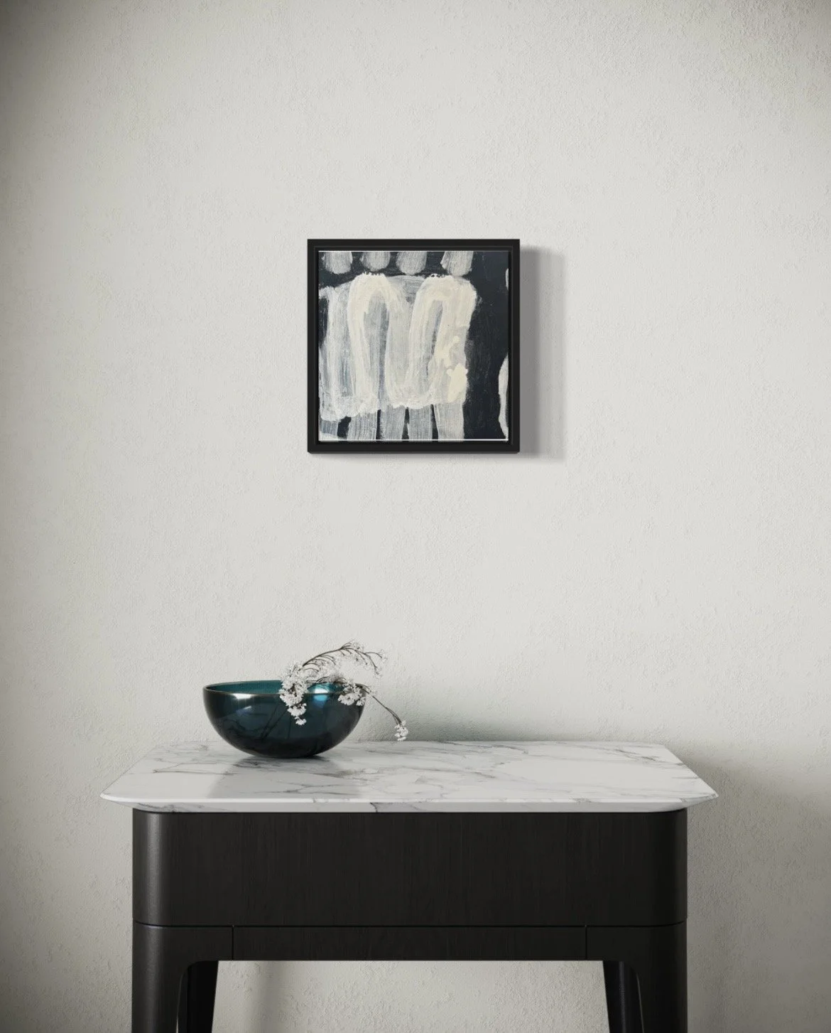 A black wooden table with a white marble top, a dark green bowl with white flowers and a branch, a white wall with a framed black-and-white painting of two abstract figures.