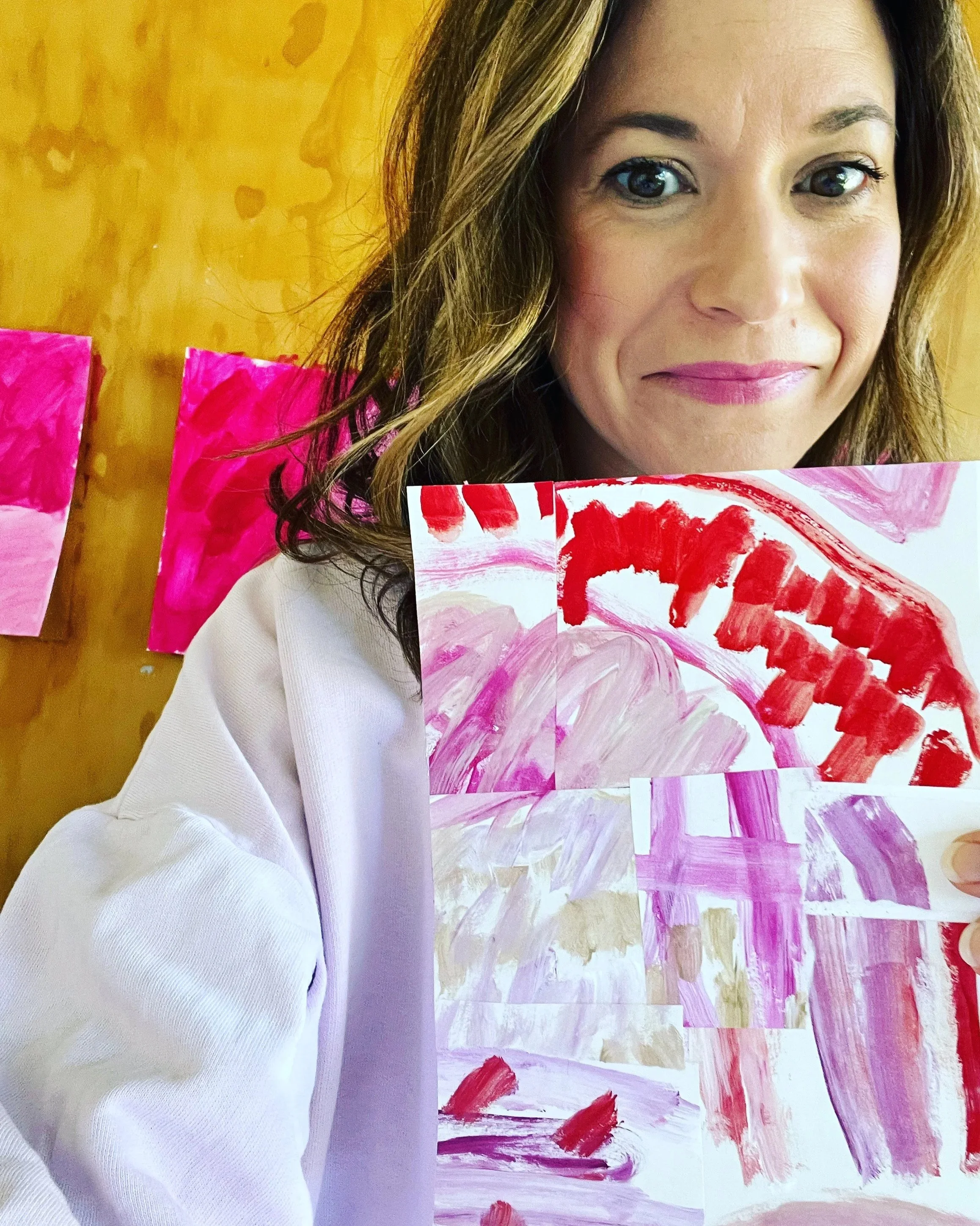 A woman with brown hair and pink lipstick is smiling at the camera. She is holding a colorful, abstract painting with pink, red, and white brushstrokes.