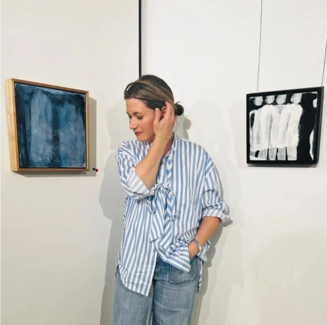 Woman wearing a striped shirt in an art gallery, standing next to abstract paintings on white walls.