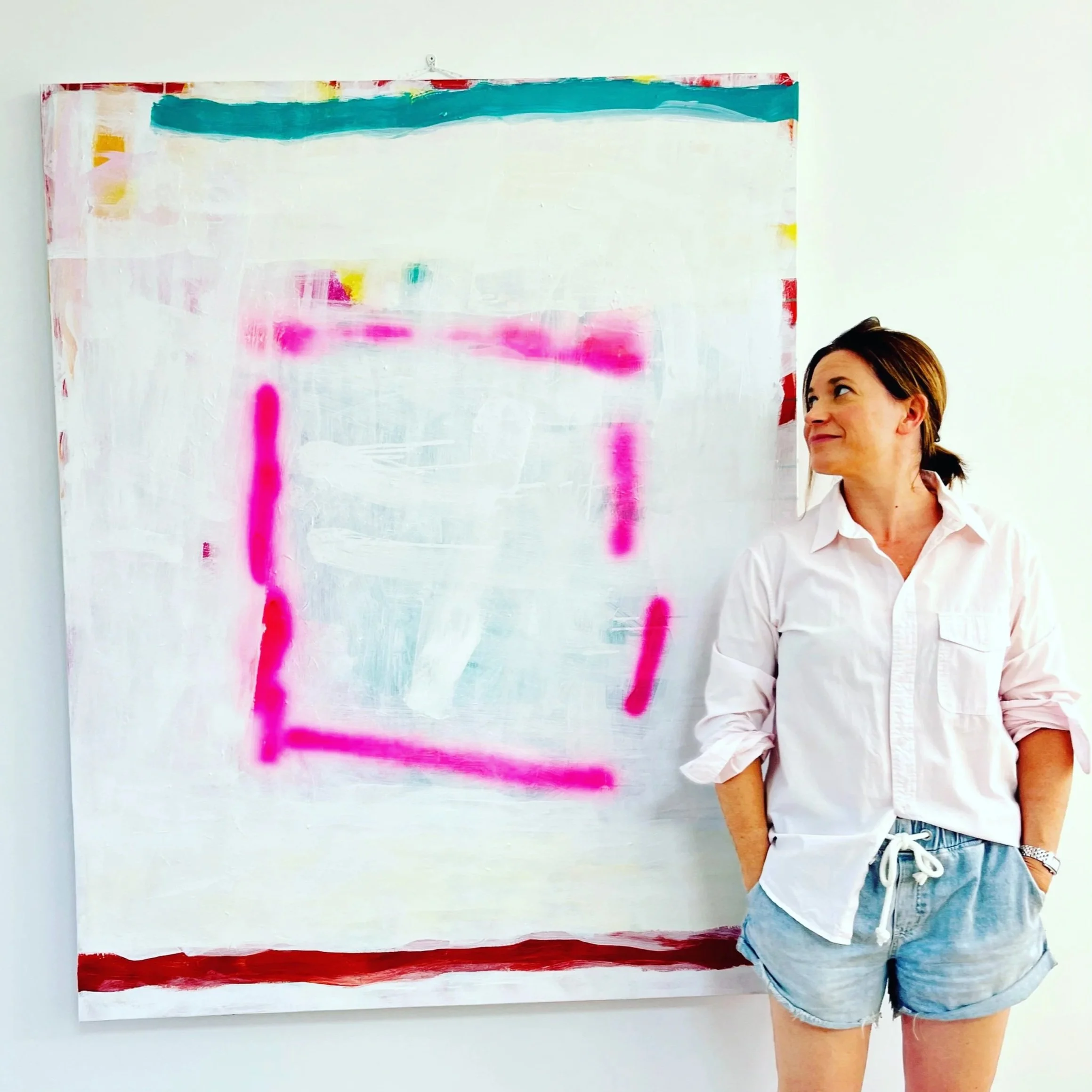 A woman standing in front of an abstract painting with horizontal blue, pink, yellow, and red streaks, wearing a white shirt and denim shorts.