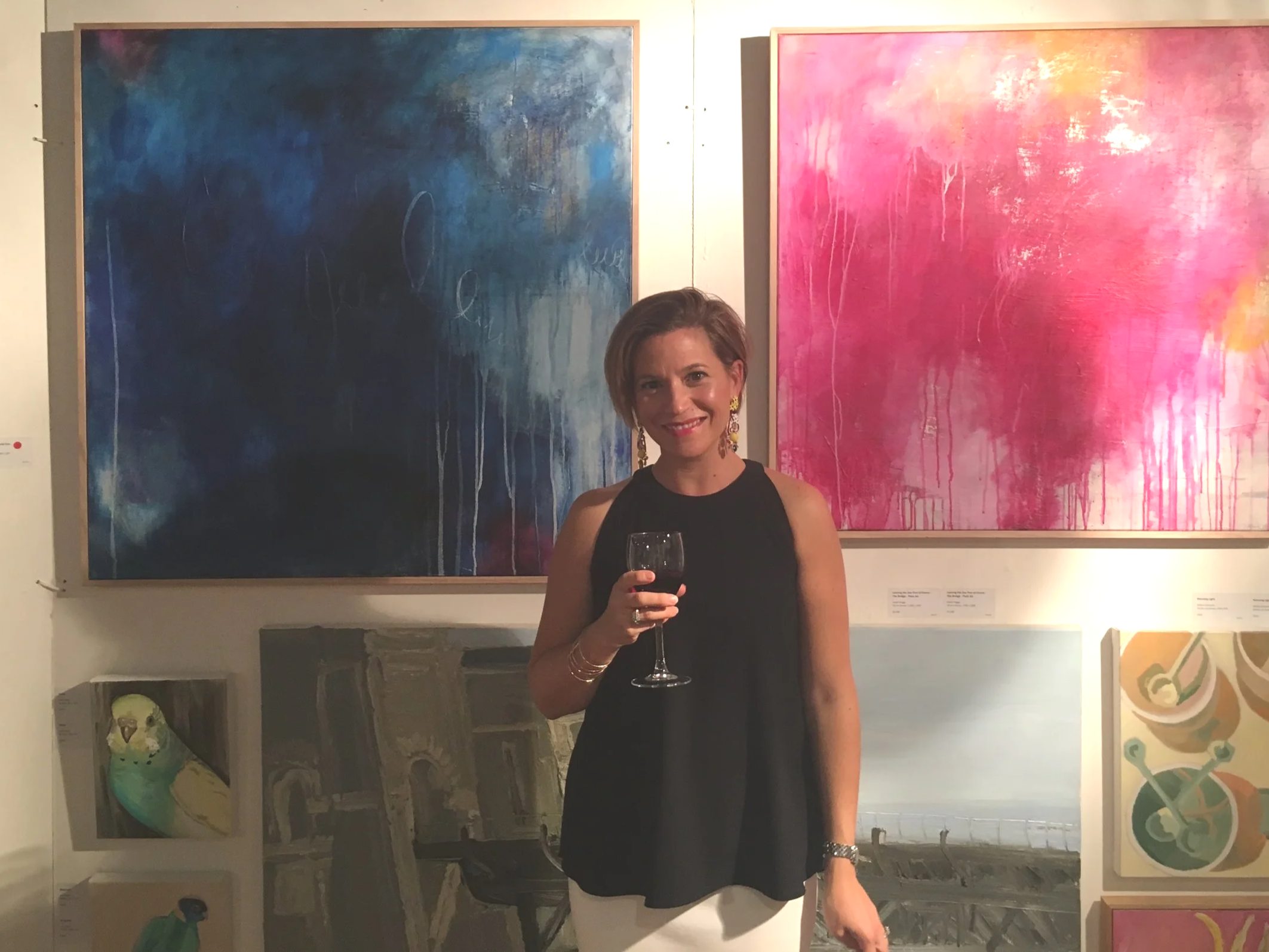 A woman in a black dress holding a glass of red wine stands in front of two large, colorful abstract paintings with shades of blue and pink, in an art gallery.
