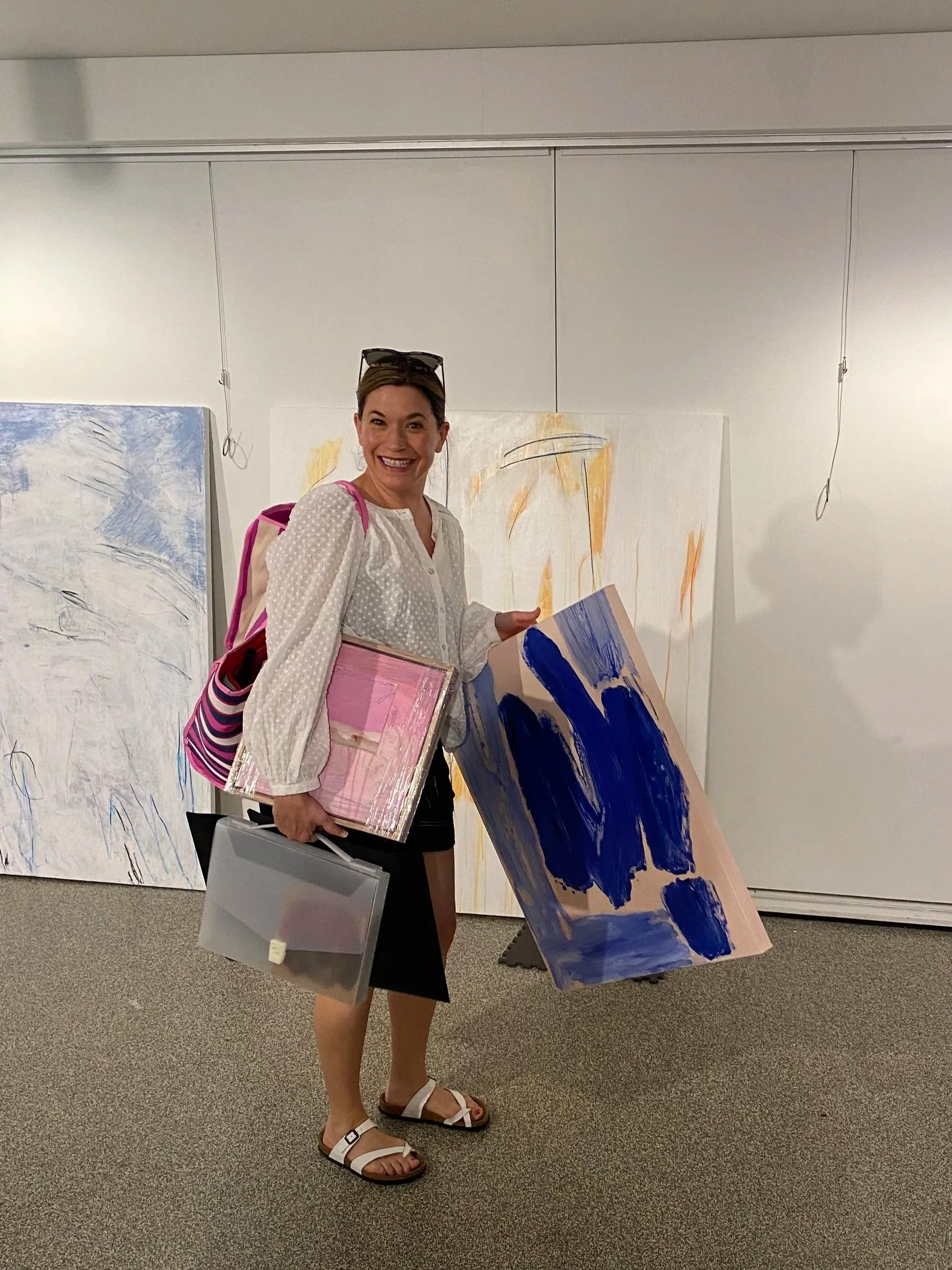A woman smiling at an art gallery holding a canvas with an abstract painting in shades of blue and pink. She is wearing a white blouse, black skirt, and white sandals, with sunglasses on her head and a pink and white striped bag.