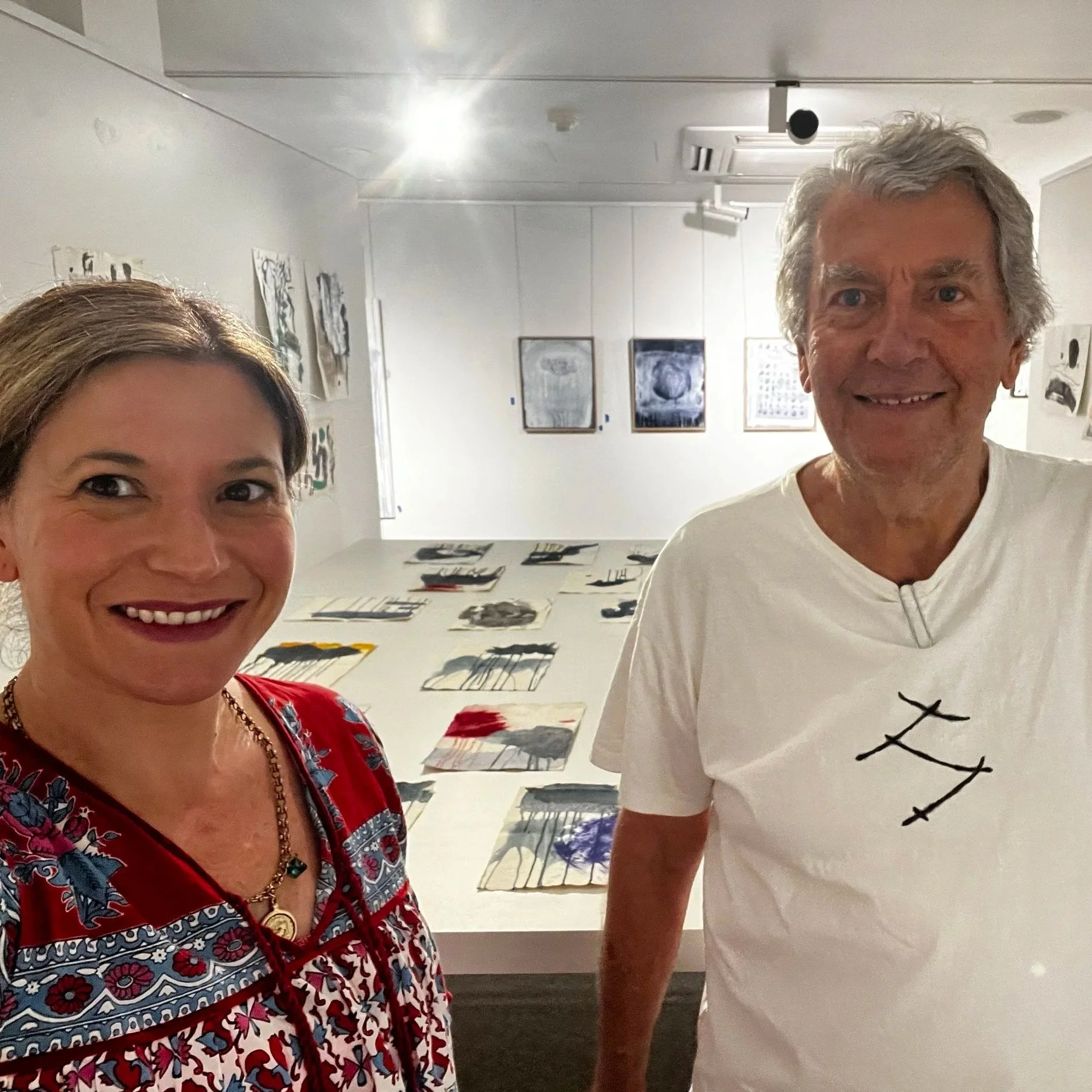 Close-up of smiling woman with short brown hair wearing a red and blue patterned dress in an art gallery with artworks displayed on white walls in the background.