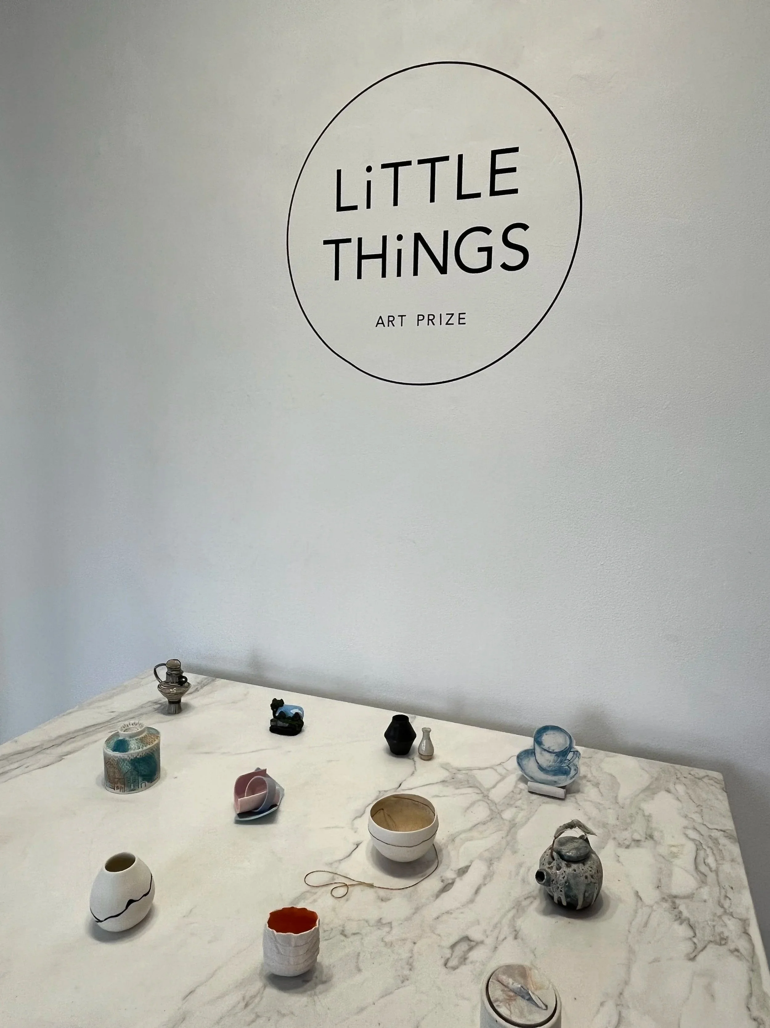 Display of various small art pieces and ceramics on a white marble table in a gallery space with a sign that reads 'Little Things Art Prize' on the wall.