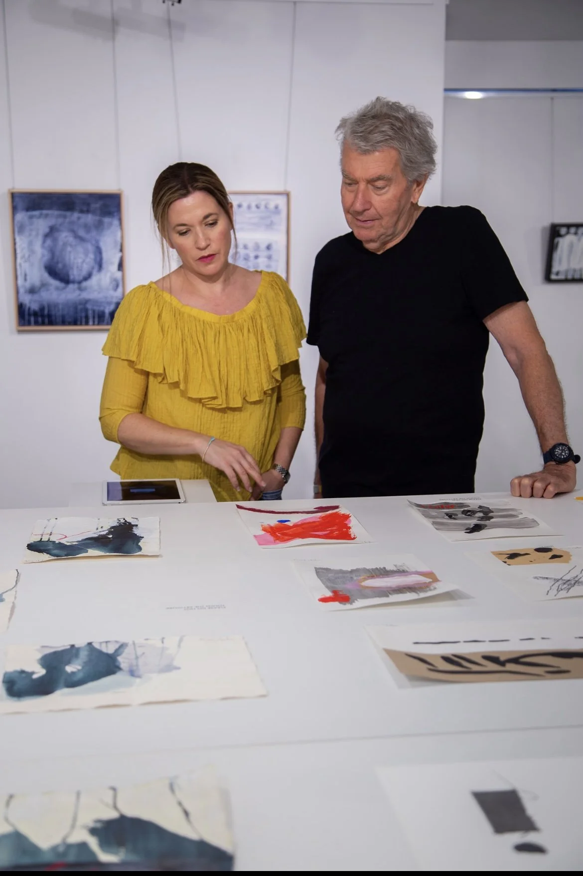 A woman in a yellow blouse and an older man in a black shirt are looking at small abstract paintings or artworks displayed on a white table. The background shows framed artwork hanging on the wall, indicating an art gallery setting.