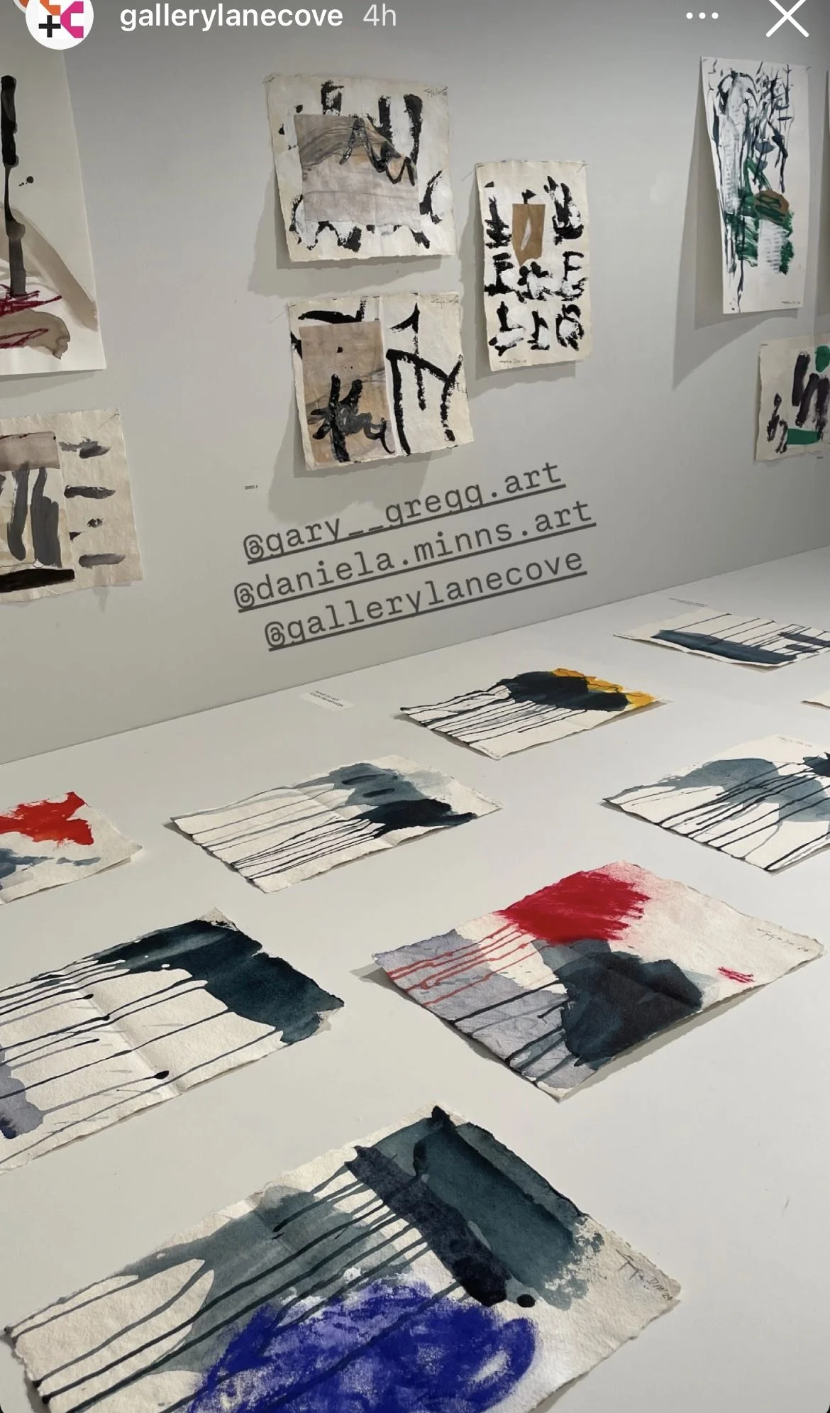 An art display featuring abstract paintings on small paper sheets and larger paintings on mounted paper. The small sheets have colorful brushstrokes and black lines, while the mounted paintings show abstract forms with black lines and various colors.