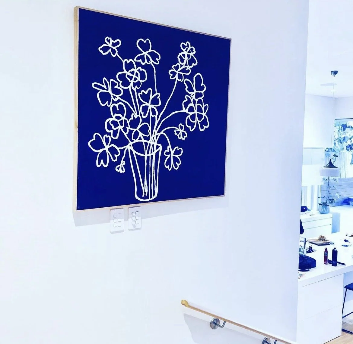 Blue artwork of a white line drawing of a vase with flowers and leaves hanging on a white wall in a modern room.