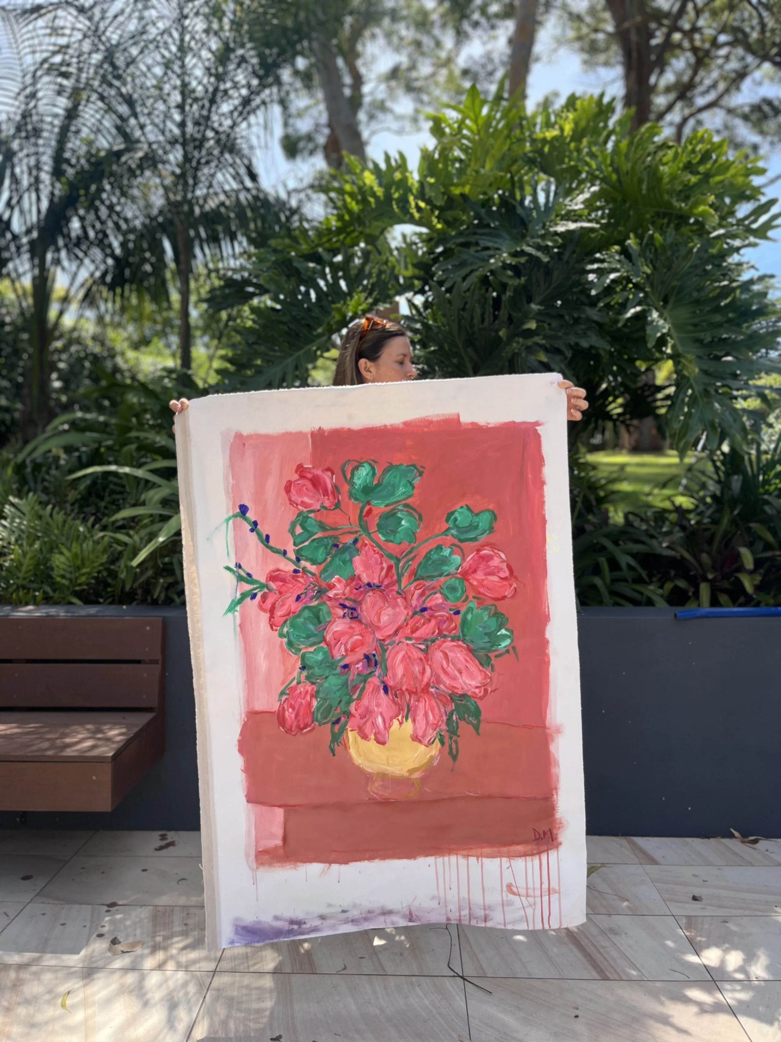 A woman holding a colorful painting of a flower vase with pink flowers against a pink background, outdoors with green trees and blue sky in the background.