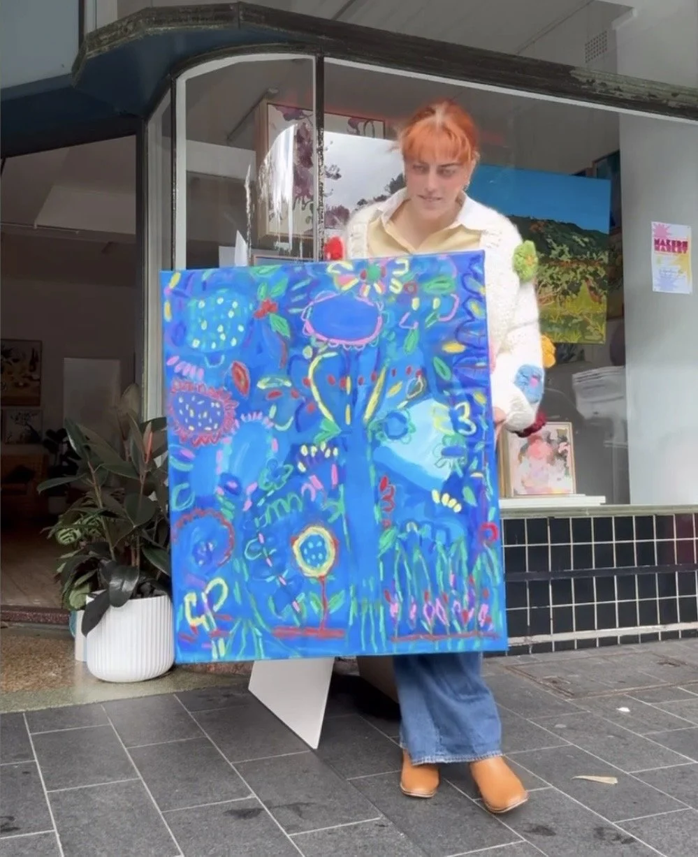 A person with short red hair wearing a white sweater and blue jeans is standing outside an art gallery, holding a colorful abstract painting with a blue background and bright, multicolored shapes and lines.