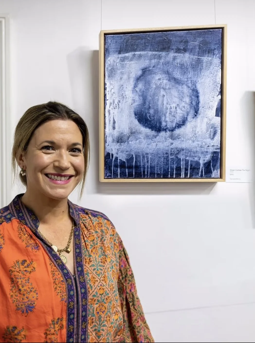 A woman with shoulder-length hair, wearing an orange and purple embroidered top, standing in front of an abstract blue painting in an art gallery.