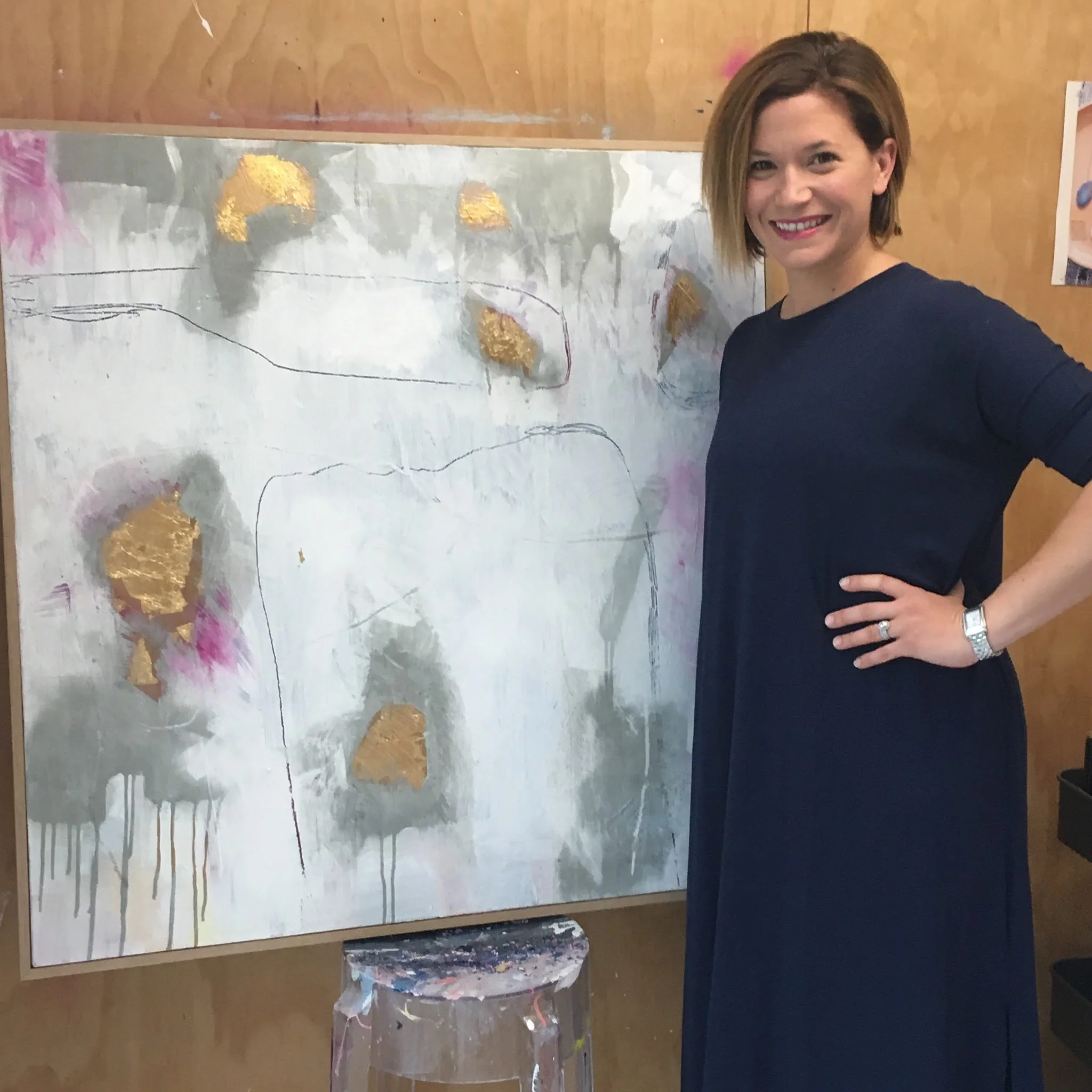 A woman with short brown hair and a dark blue shirt standing next to a large abstract painting with white, gold, and pink colors on a wooden wall.