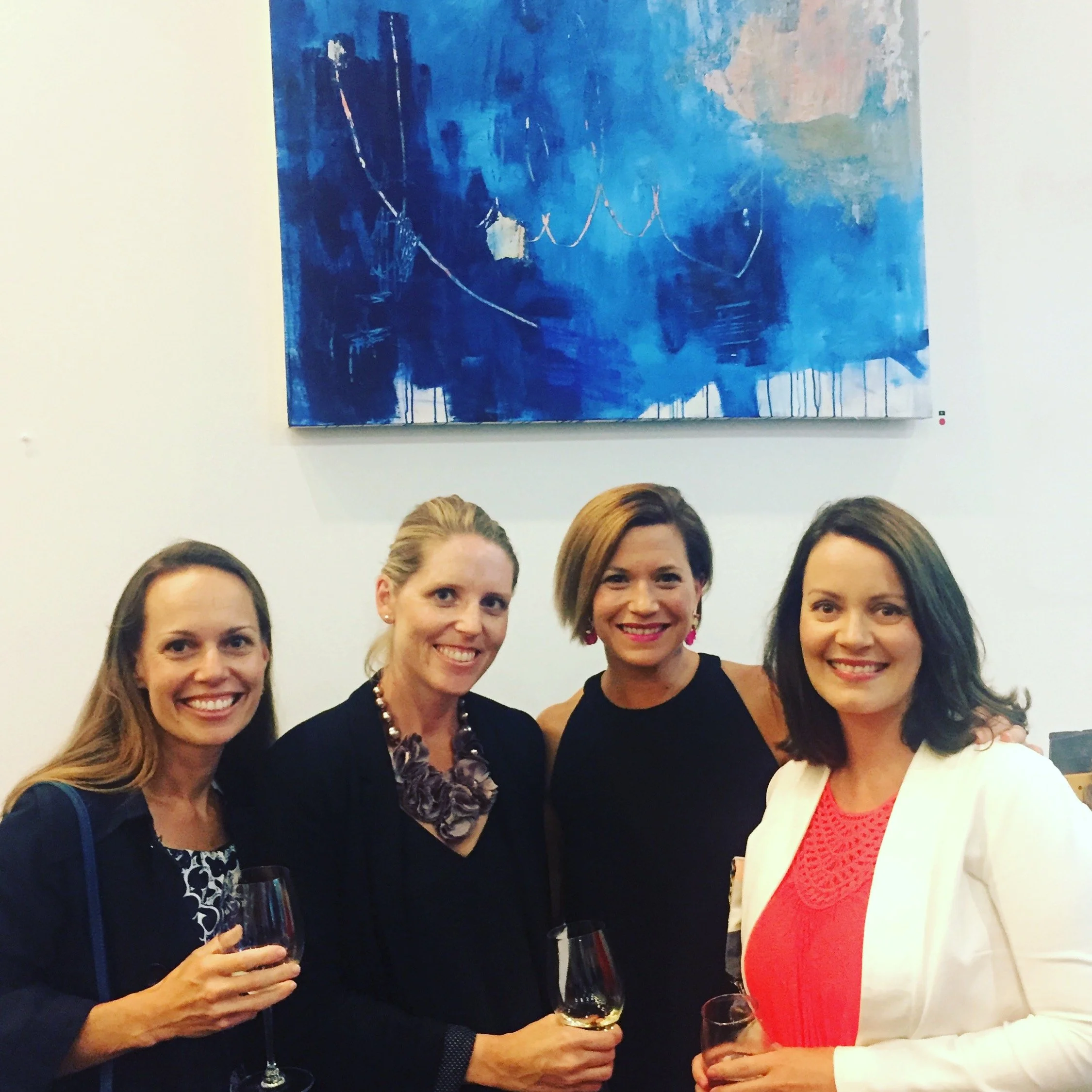 A group of women at a social gathering holding wine glasses and smiling, with a blue abstract painting on the wall behind them.