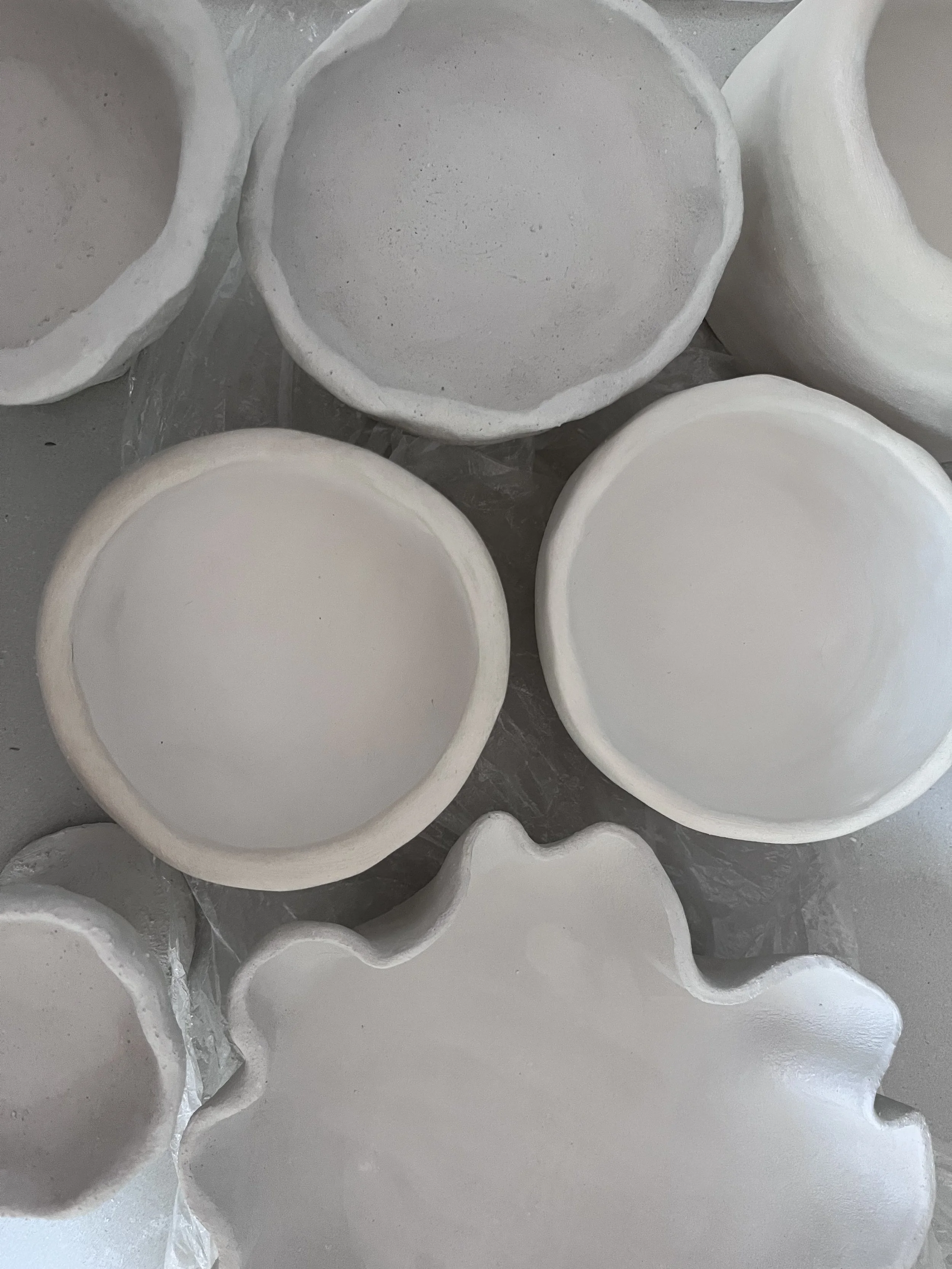 Multiple white ceramic bowls, some with uneven edges, stacked and arranged on a surface, with some partially covered in plastic wrap.