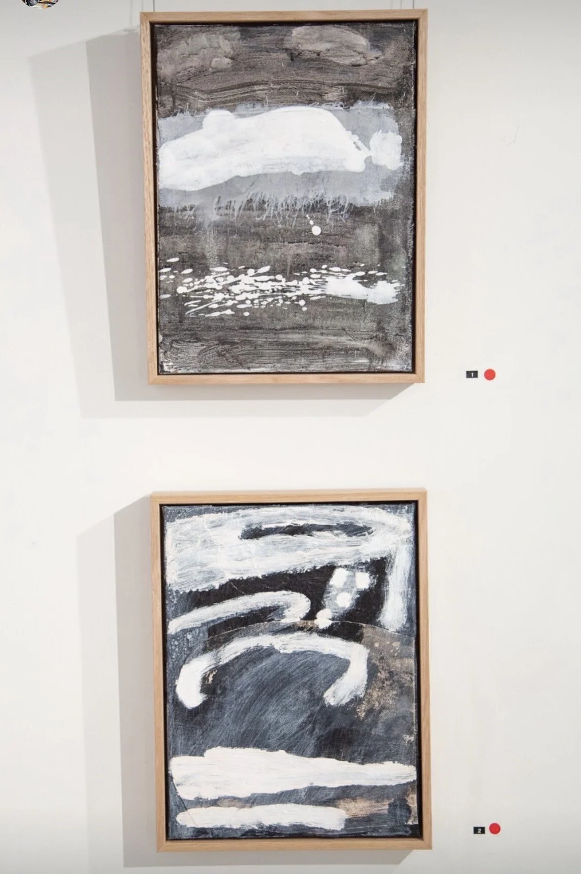 Two abstract black and white paintings framed in wood, displayed on a white wall in an art gallery. The top painting features a cloud-like shape with water reflections, while the bottom painting shows bold, swirling brushstrokes.