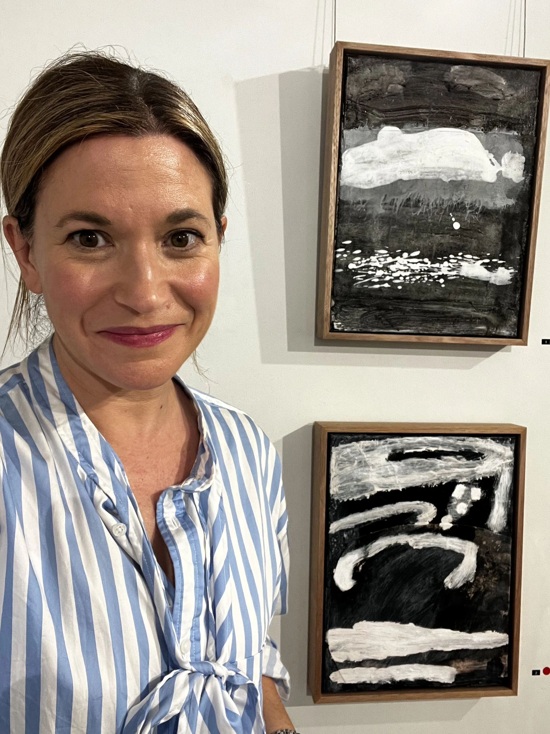 A woman wearing a blue and white striped shirt standing in front of modern black and white abstract paintings on a white gallery wall.