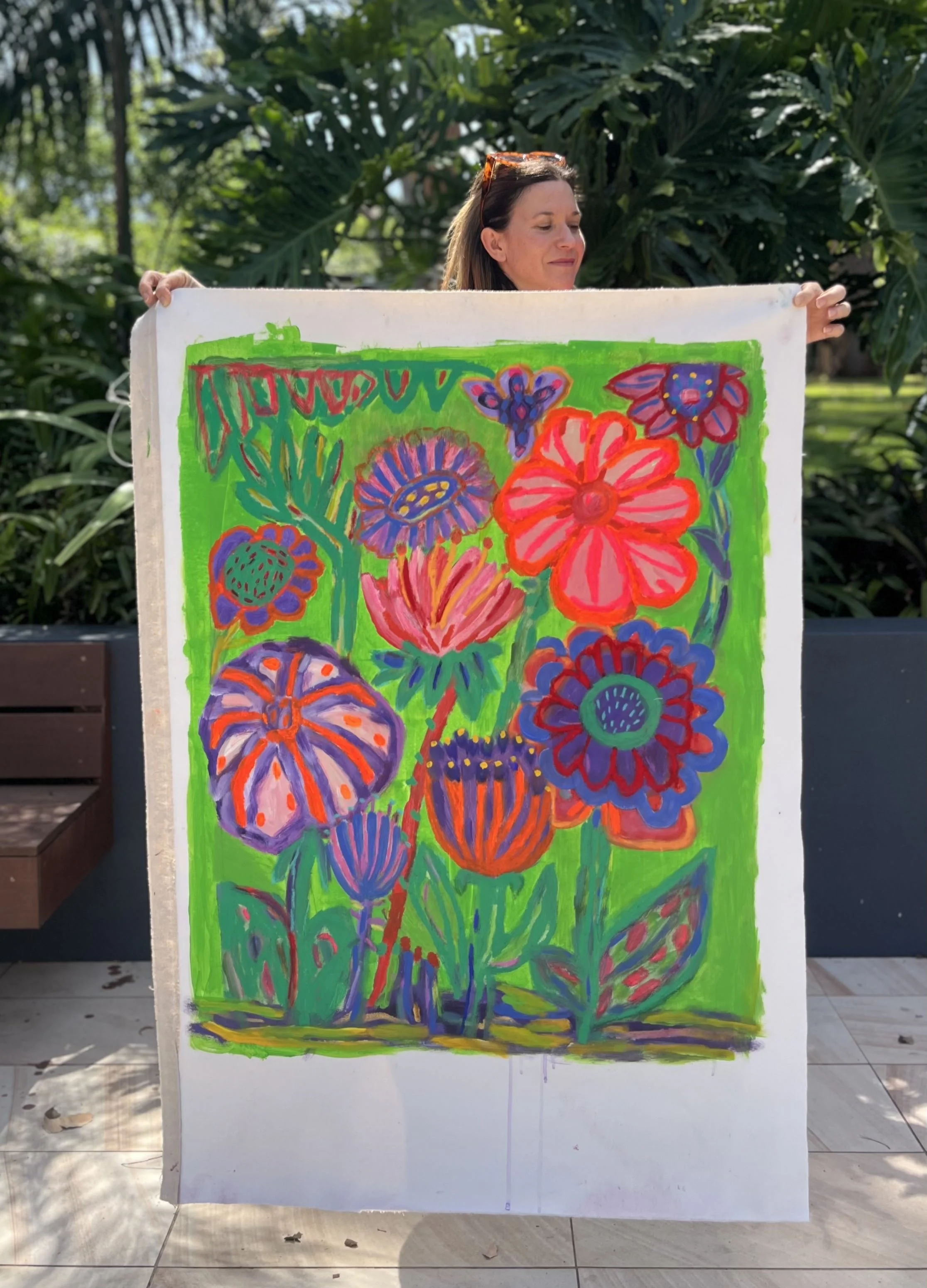 Colorful floral painting on a white canvas held by a woman outdoors with green foliage in the background.