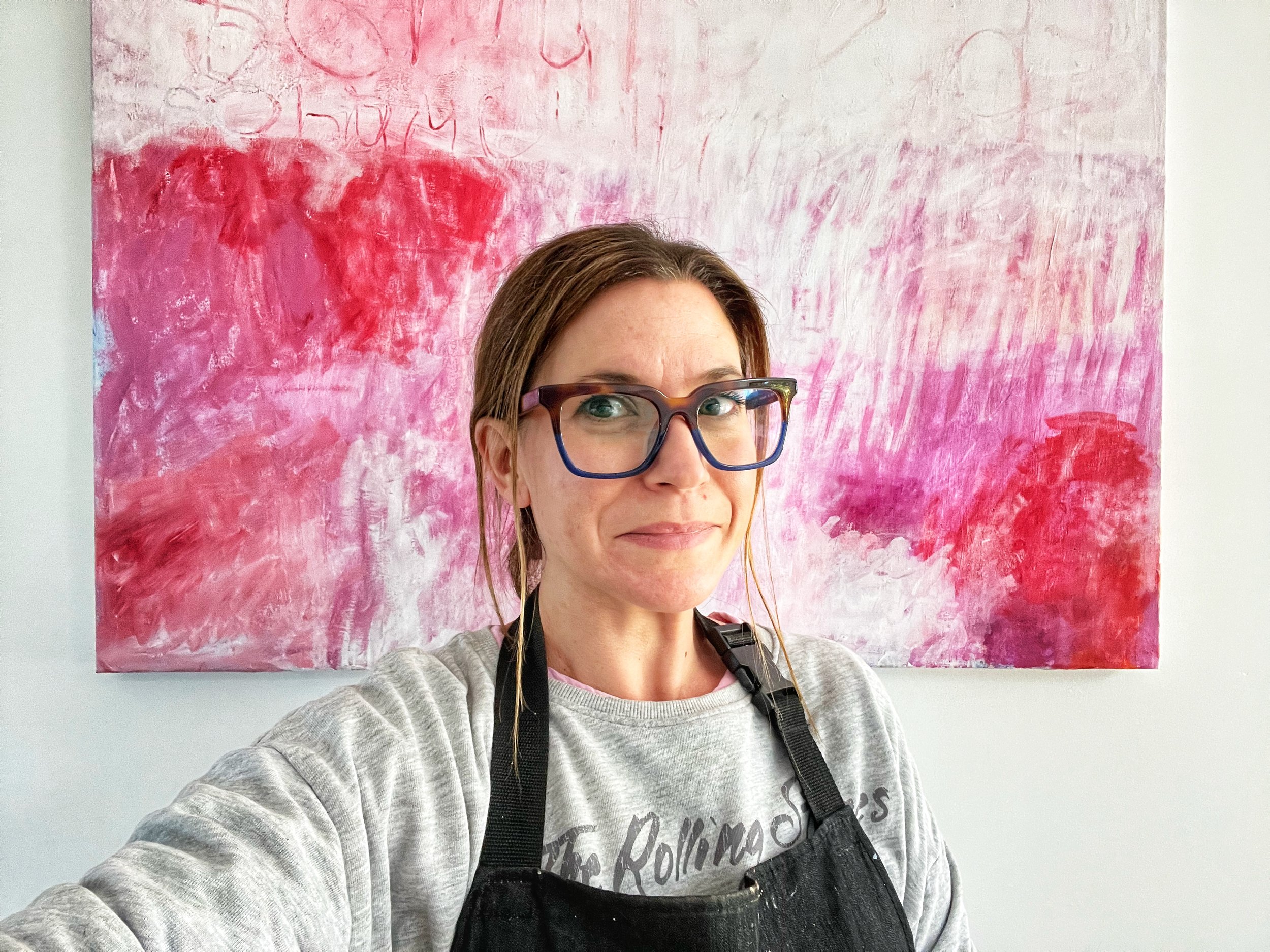 A woman with brown hair, wearing glasses, a gray sweatshirt, and an apron, taking a selfie in front of an abstract pink and red painting.